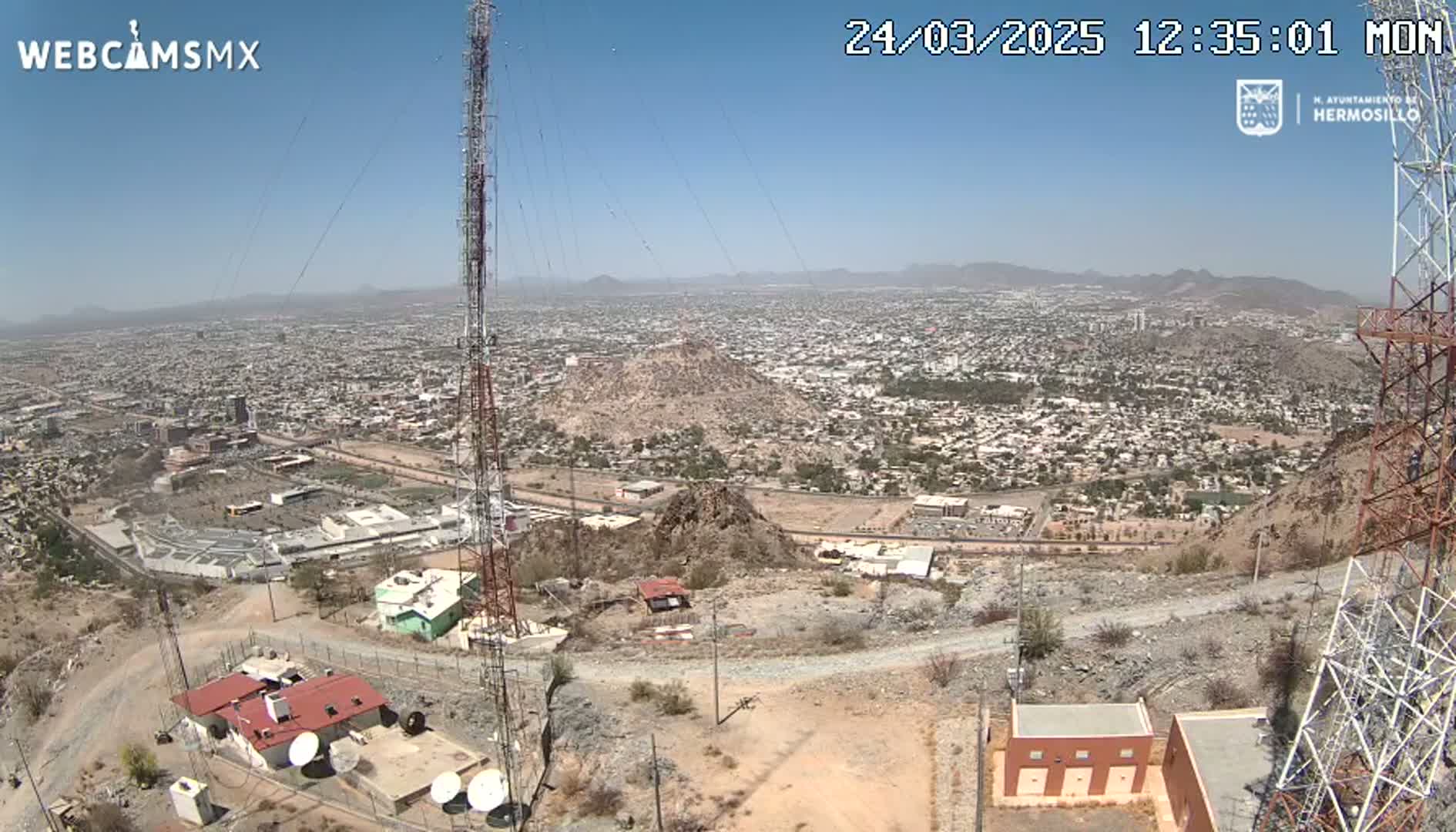 A sprawling city is visible in the distance under a clear, sunny sky, viewed from a rocky, high vantage point with communication towers and small buildings in the foreground.