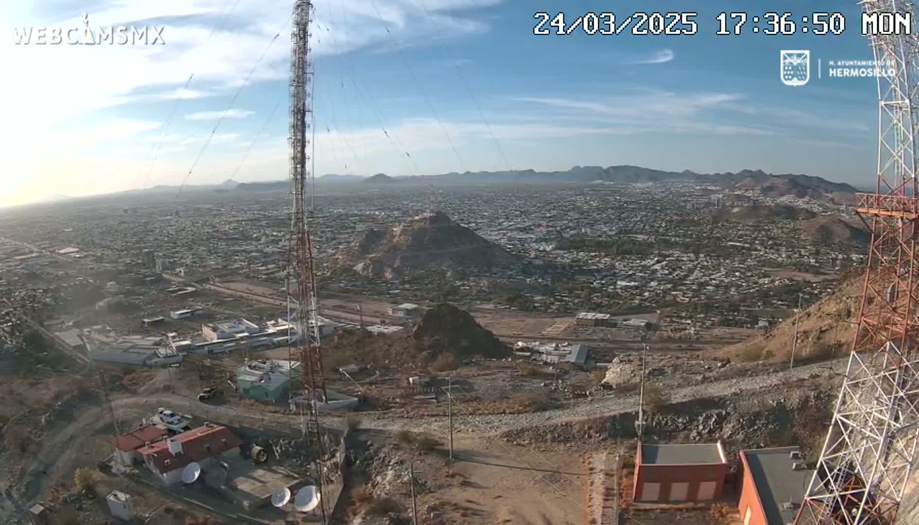 A high-angle, long shot on a clear day shows a large city sprawling across a valley, with a rocky hill in the foreground featuring communication towers and buildings.