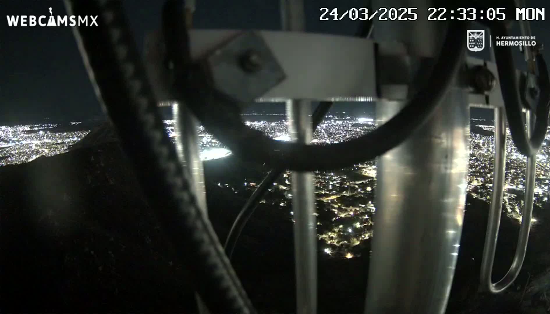 A nighttime view from a high vantage point shows a sprawling city illuminated below, seen through the metal framework of a structure.