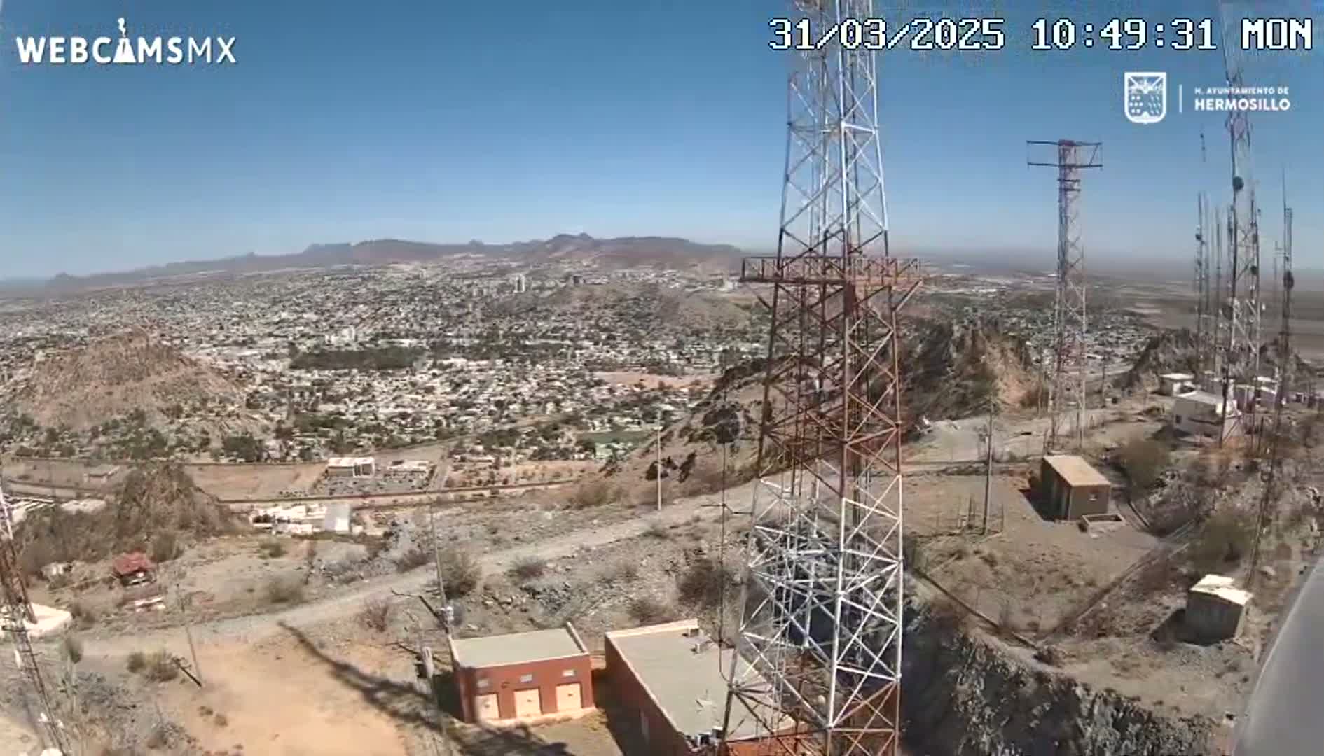 A high-angle view on a clear, sunny day shows several communication towers atop a rocky hill overlooking a sprawling city and distant mountains.