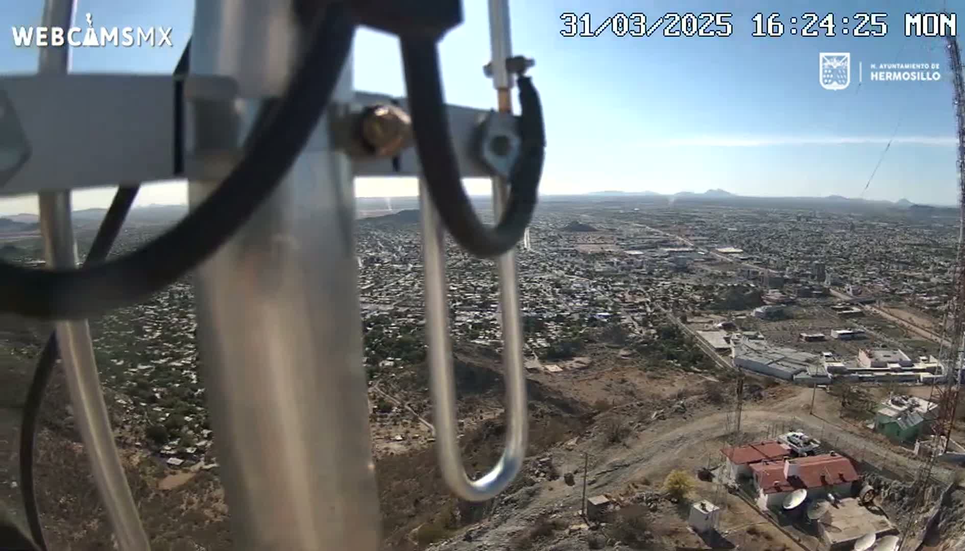 A high-angle view from a tower shows a sprawling city under a clear, sunny sky, with a small building and surrounding structures visible in the foreground.