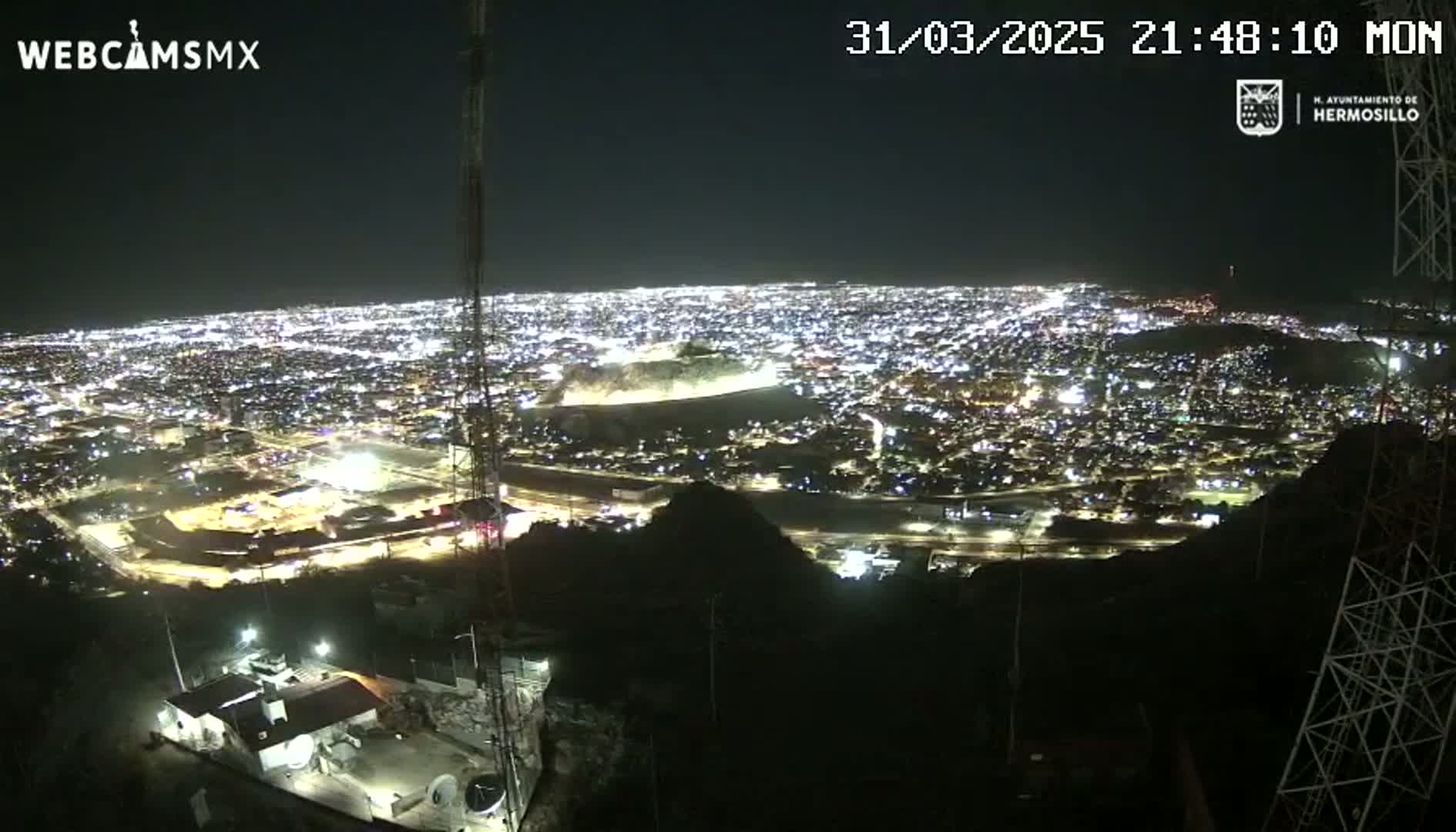 A nighttime, high-angle view shows a sprawling city illuminated by numerous lights, nestled among dark hills.