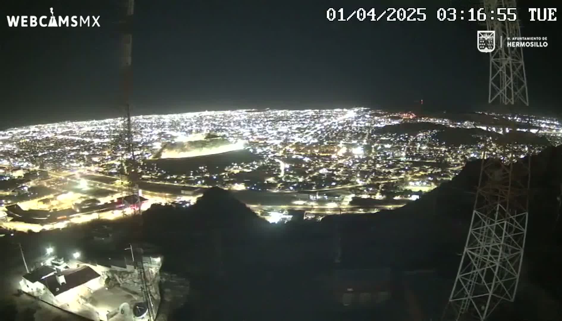 A nighttime, high-angle view shows a sprawling city illuminated with countless lights, as seen from a hilltop.