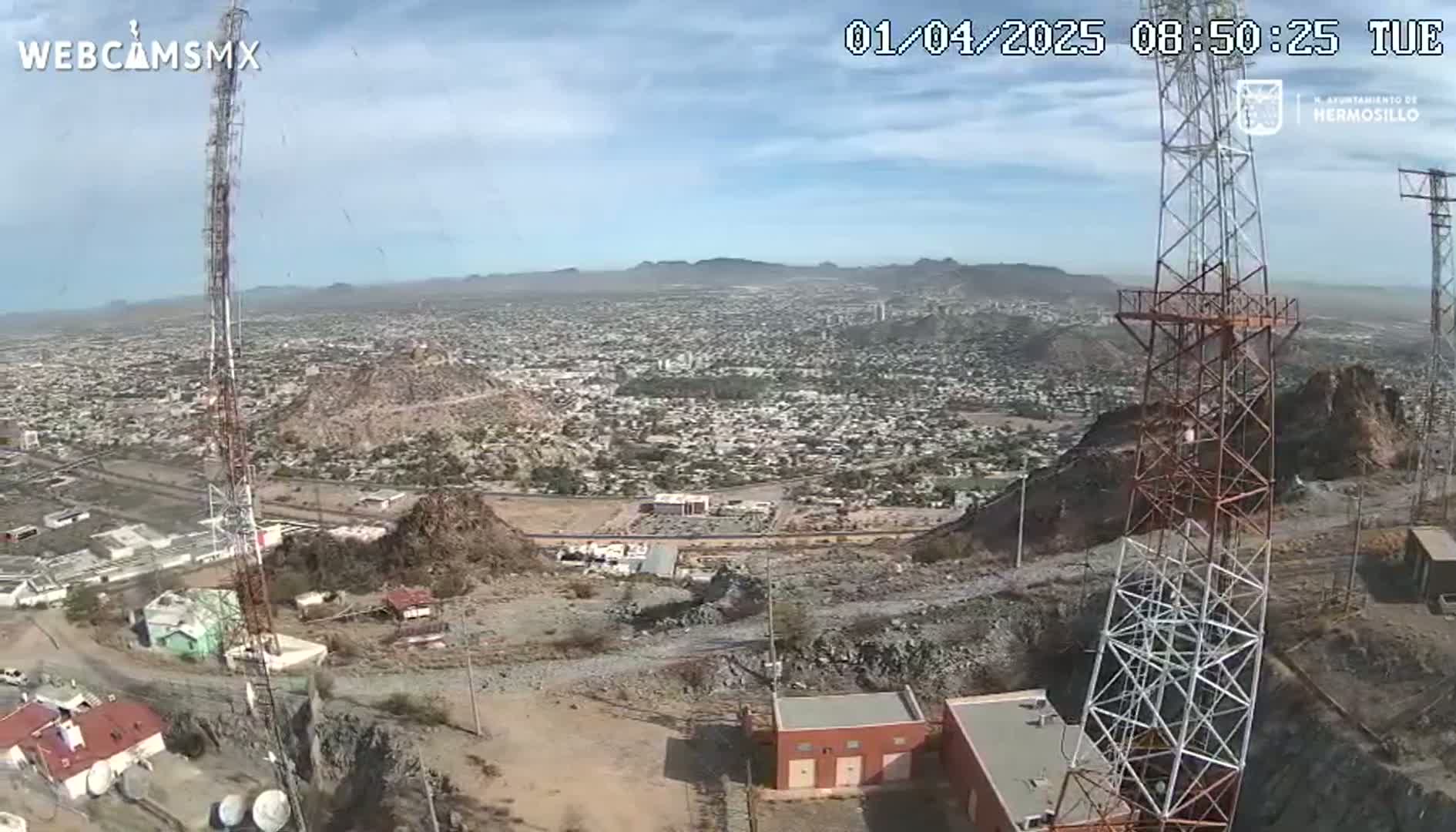 A high-angle, long shot on a clear day shows a sprawling city nestled in a mountainous region, with two tall communication towers prominently featured in the foreground.