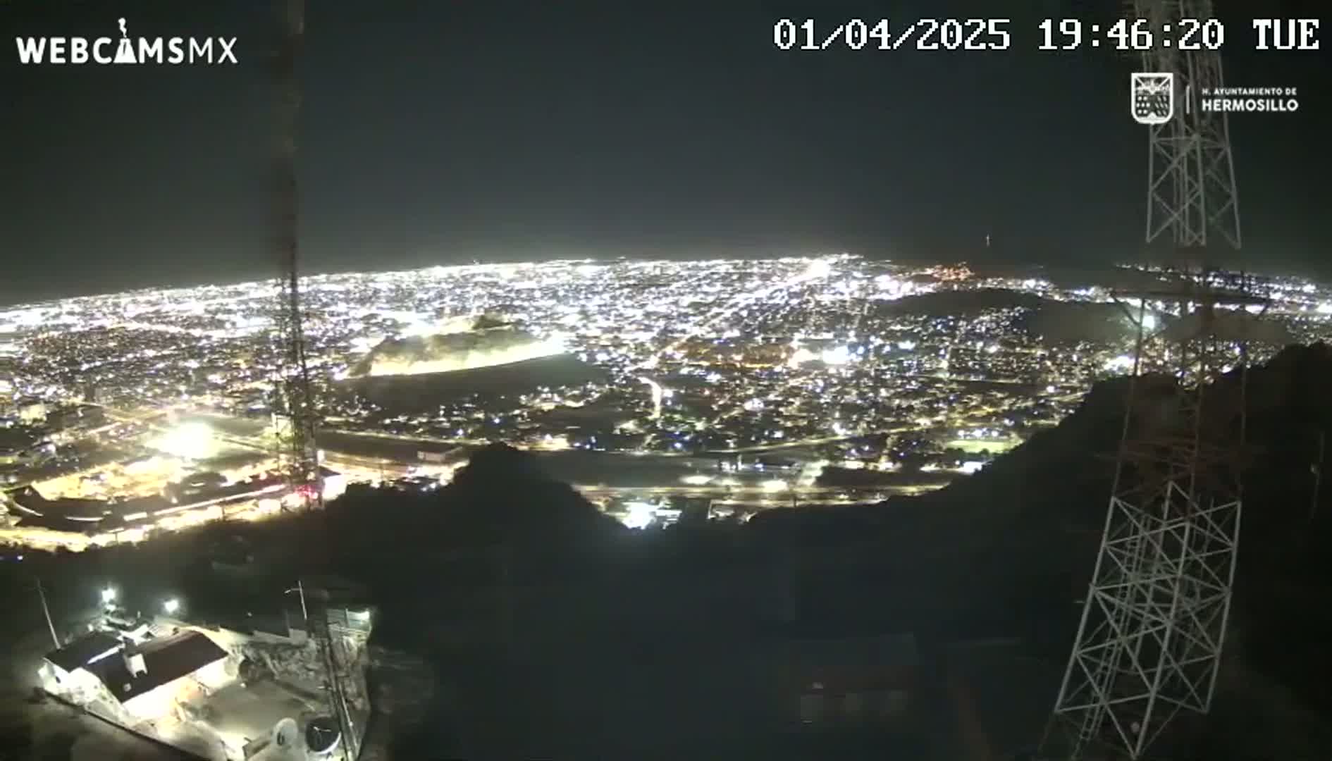 A nighttime, high-angle view of a brightly lit city sprawling across a valley, seen from a vantage point on a hill, with a tall communication tower in the foreground.