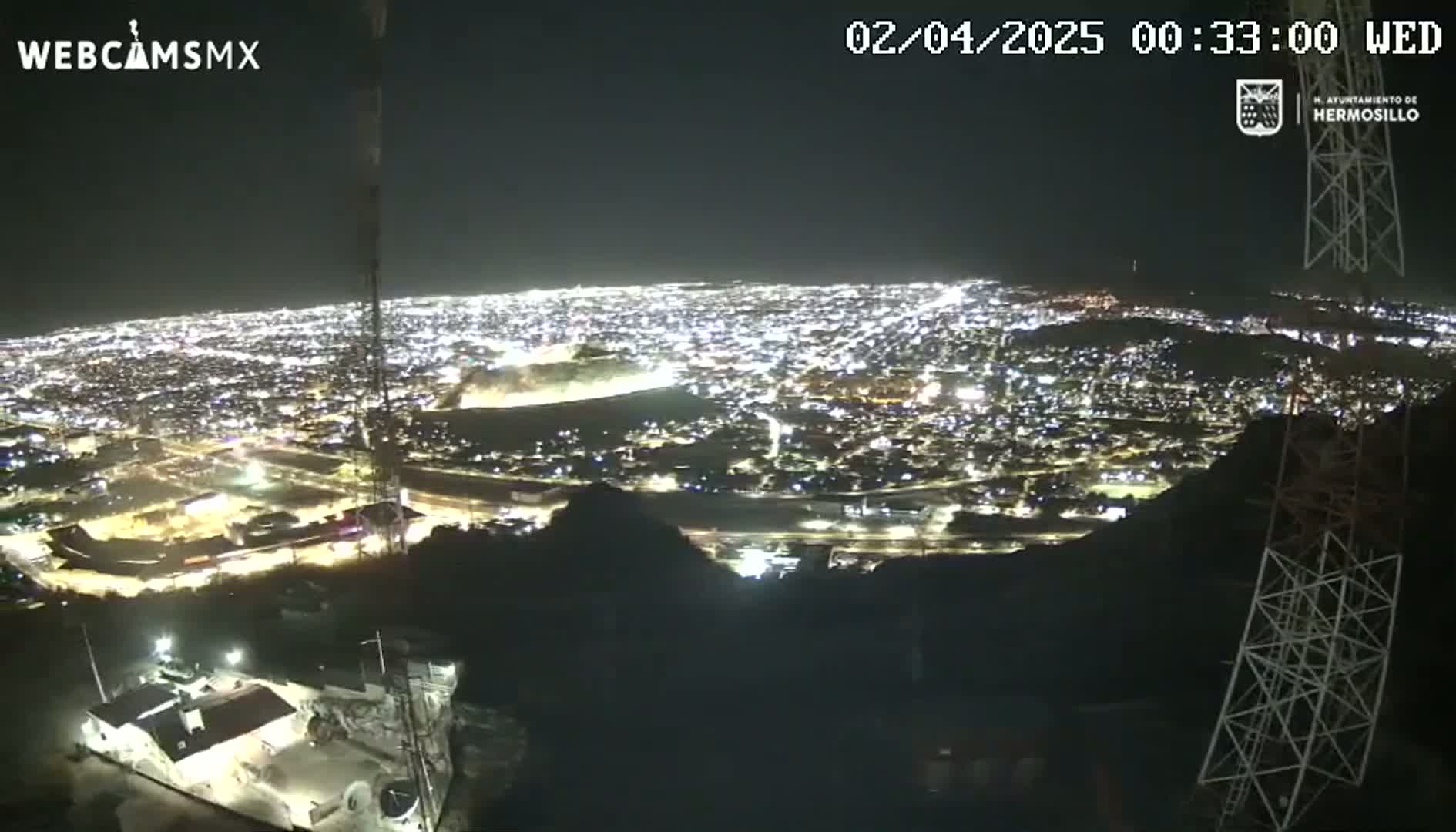 A nighttime, high-angle view shows a sprawling city illuminated by countless lights, seen from a vantage point on a hill or mountain.