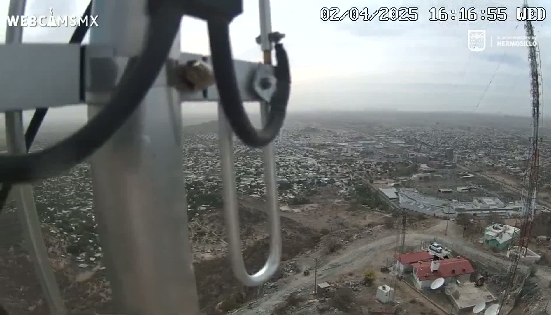 A hazy, overcast view of a sprawling city is seen from a high vantage point, partially obscured by a metal structure.