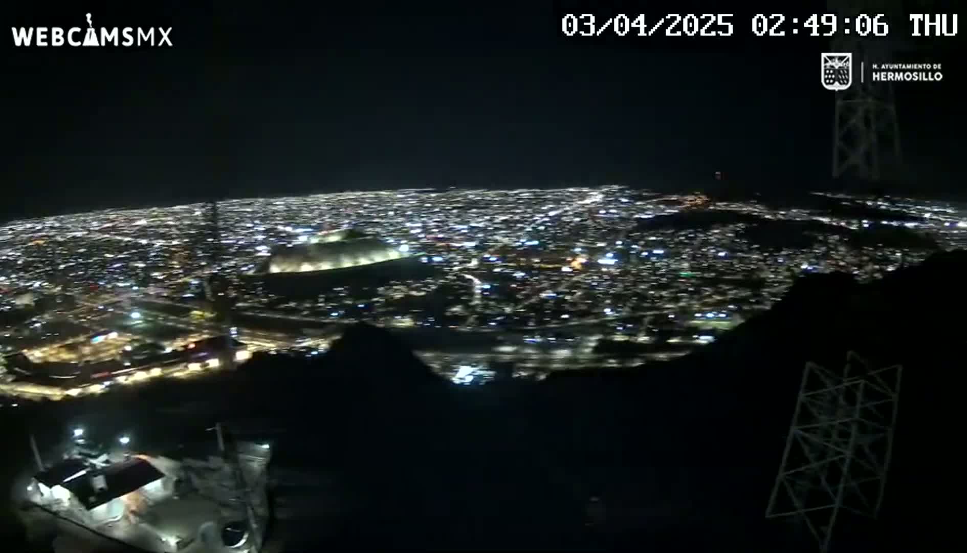 A nighttime, high-angle view shows a sprawling city illuminated below, seen from a dark, mountainous vantage point.