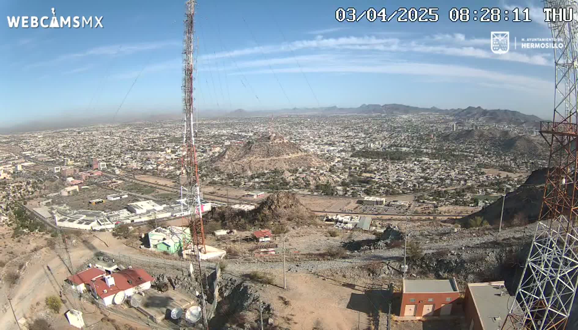 A high-angle, long shot on a clear, sunny day shows a sprawling city nestled in a mountainous region, with a tall communication tower and various buildings visible in the foreground.