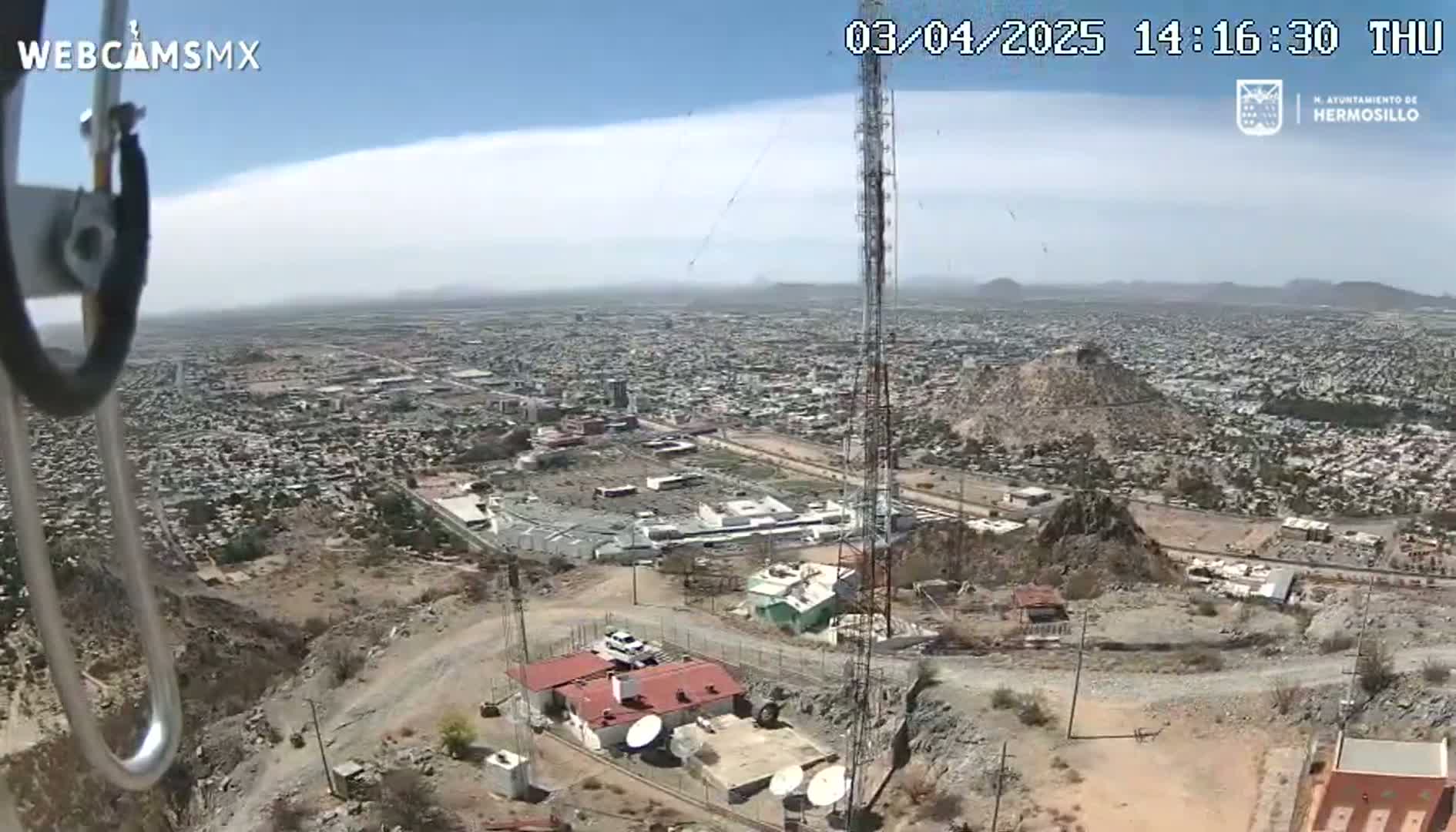 A high-angle, long shot on a sunny day shows a sprawling city below a rocky hillside with a communications tower and small buildings, viewed from what appears to be a camera mounted on a structure.