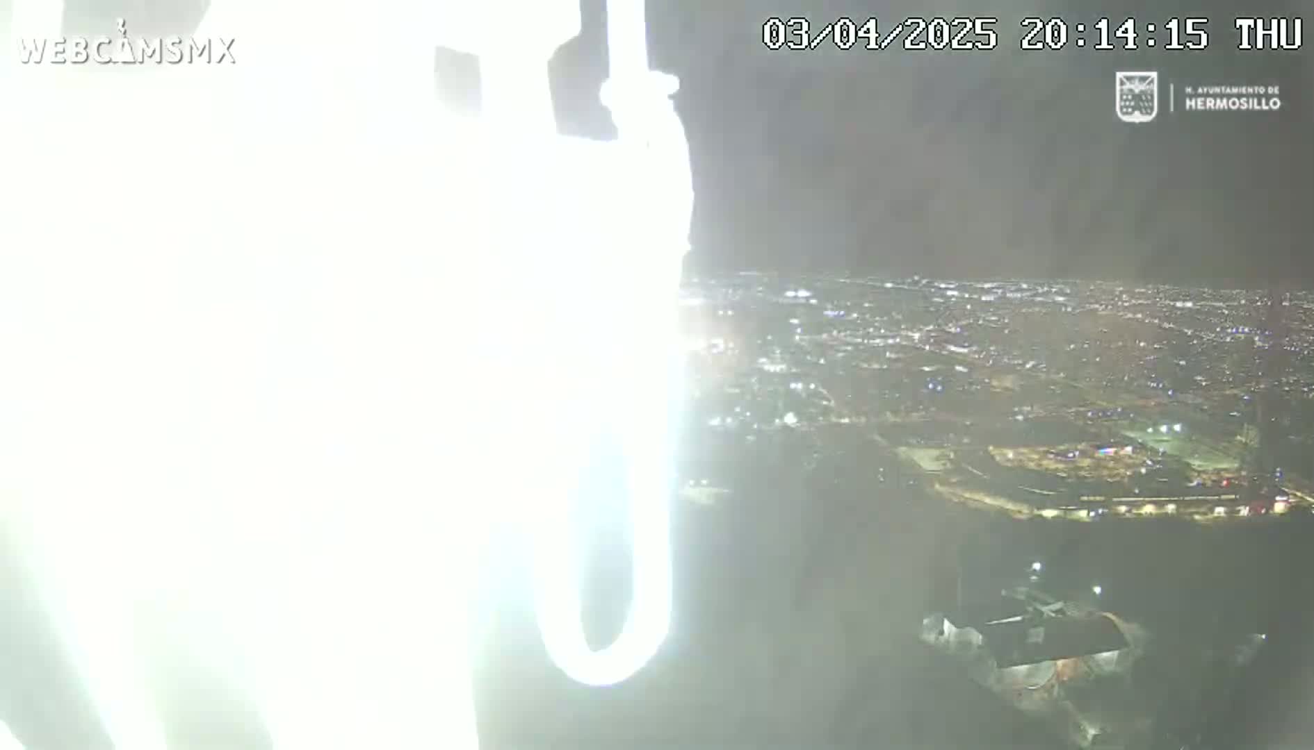 A brightly lit object obscures the view of a nighttime city scene under hazy conditions.