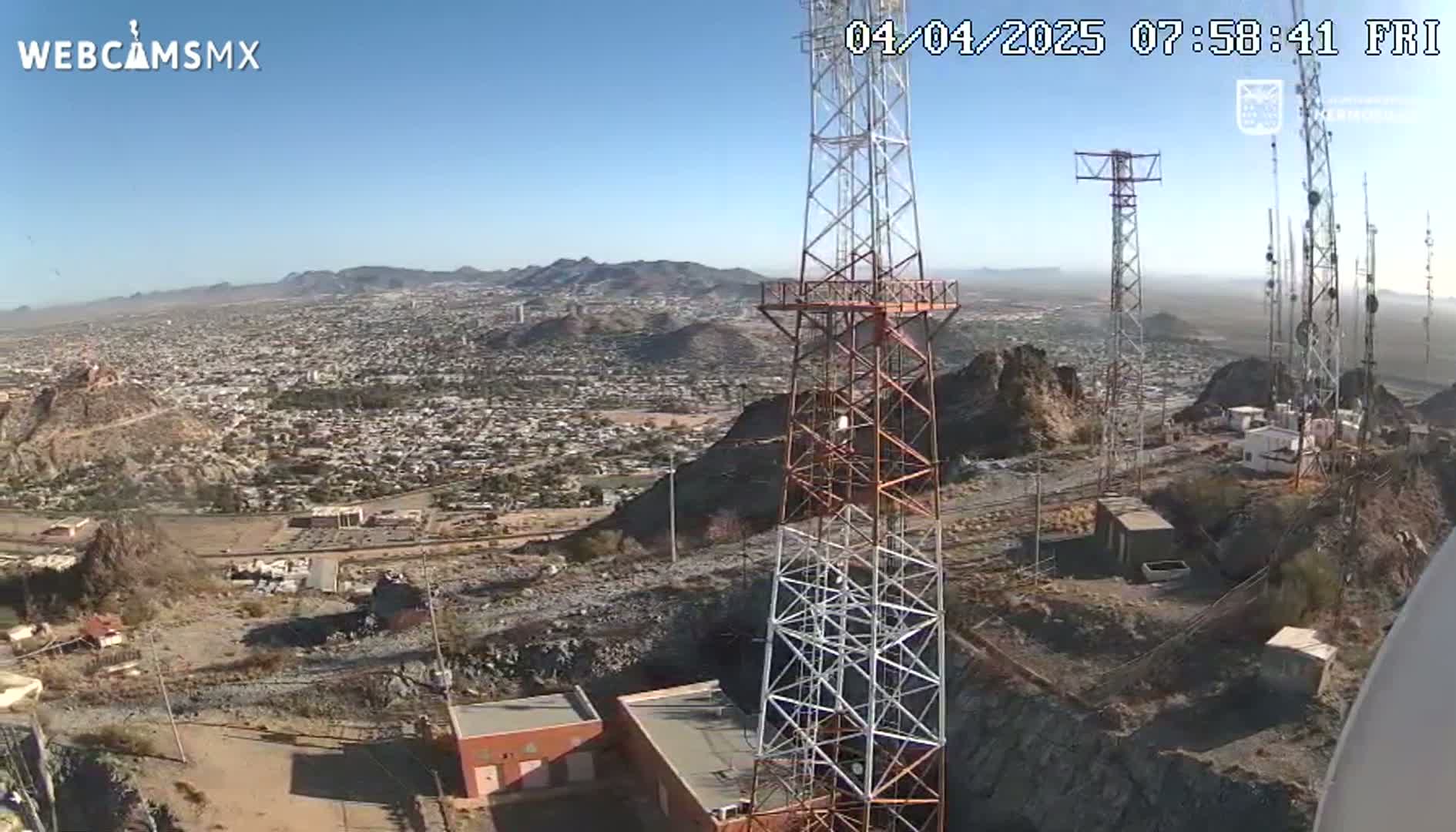 A high-angle, long shot on a clear, sunny day shows several communication towers atop a rocky hill overlooking a sprawling city and distant mountains.