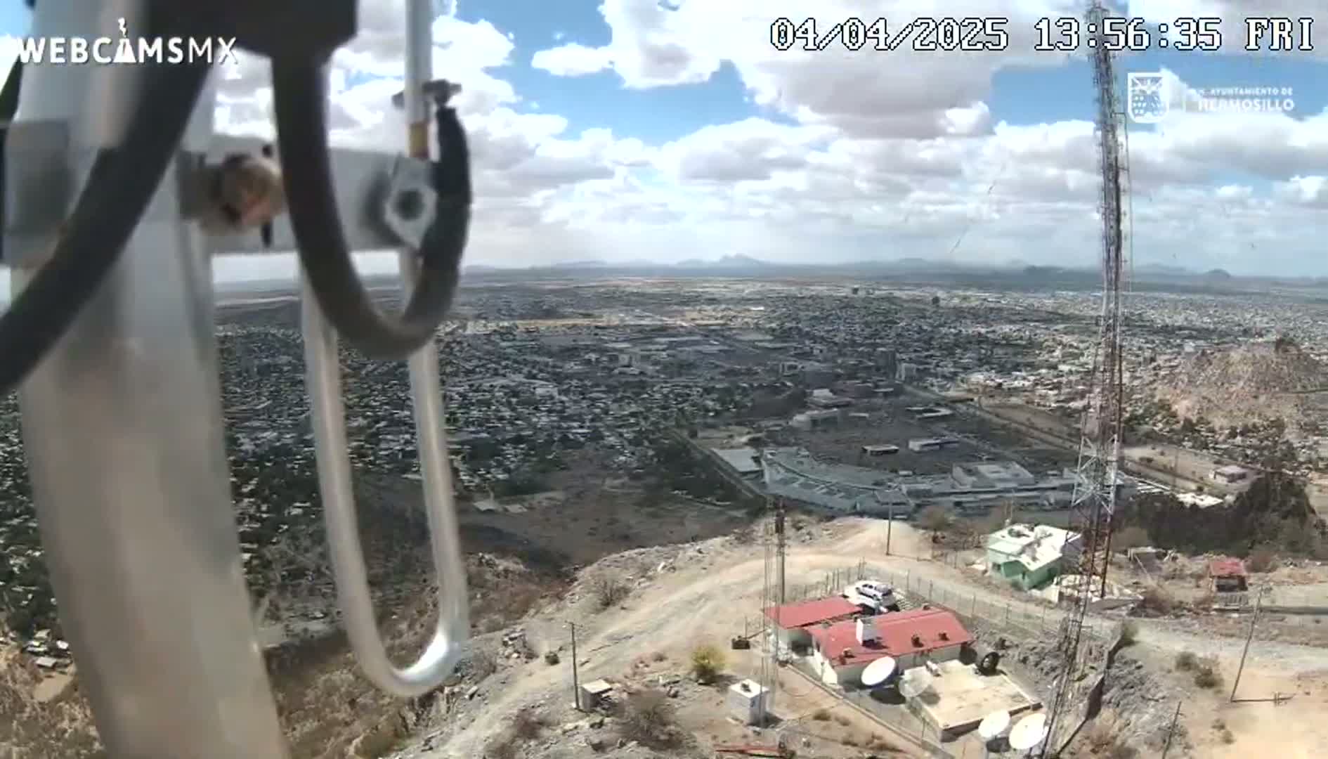 A high-angle view shows a sprawling city under partly cloudy skies, seen from a hilltop where a small building and tall communication tower are located.