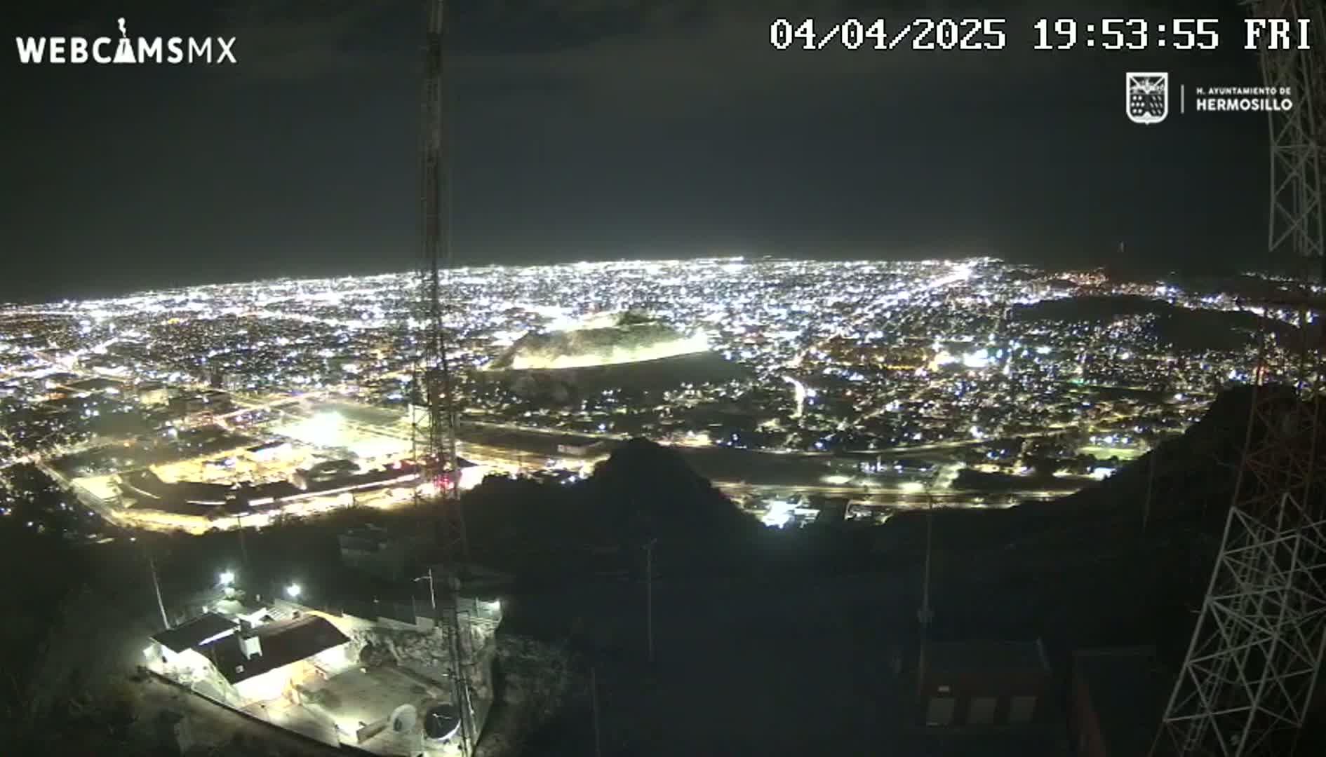 A nighttime view from a hilltop shows a sprawling city illuminated below, under a clear sky.
