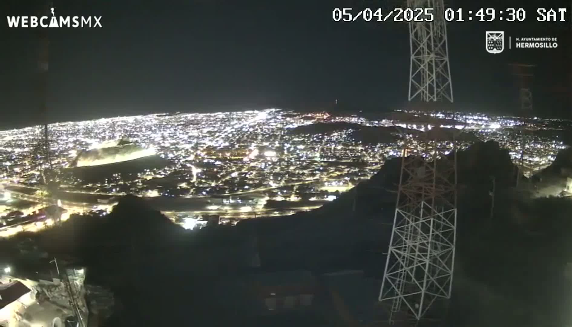 A nighttime view from a hilltop shows a sprawling city illuminated below, with several tall communication towers in the foreground under a clear, dark sky.