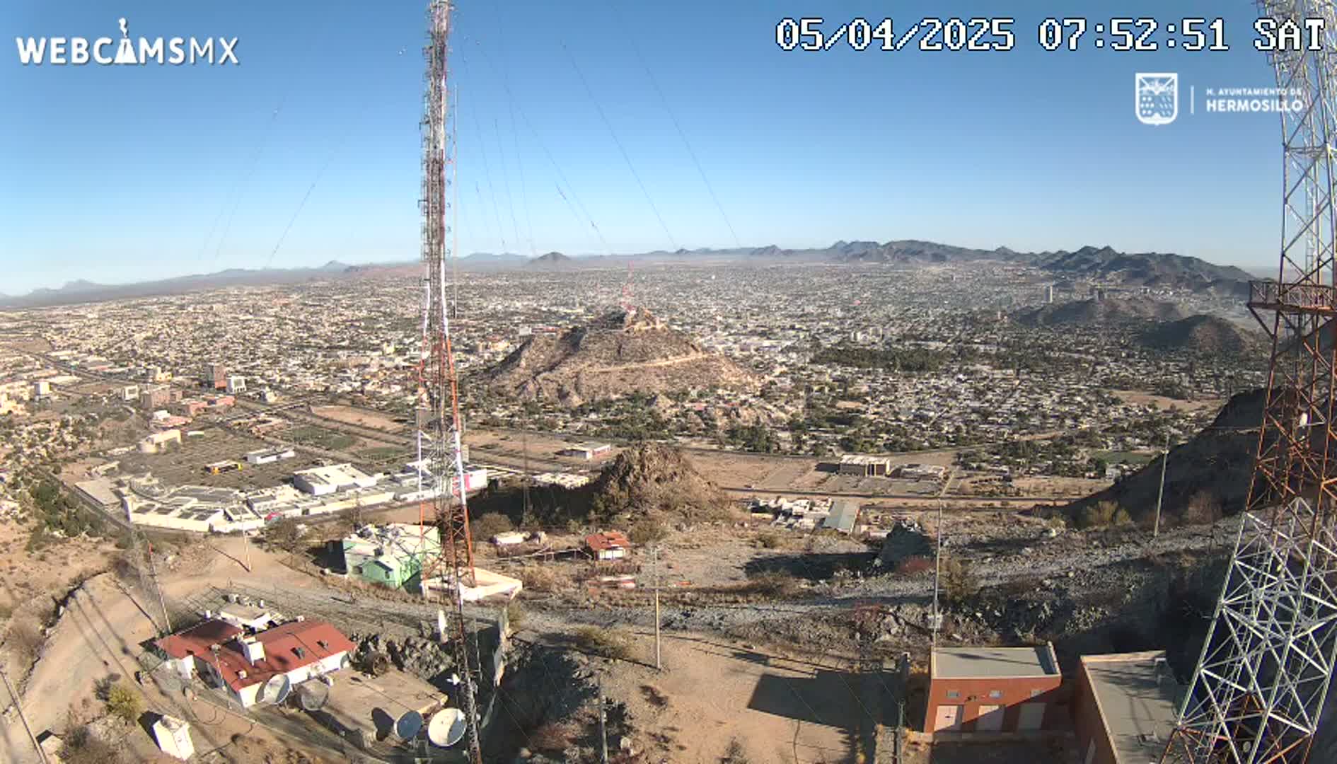 A high-angle, long shot on a clear day shows a city sprawling across a valley, viewed from a rocky hillside with communication towers and buildings nearby.