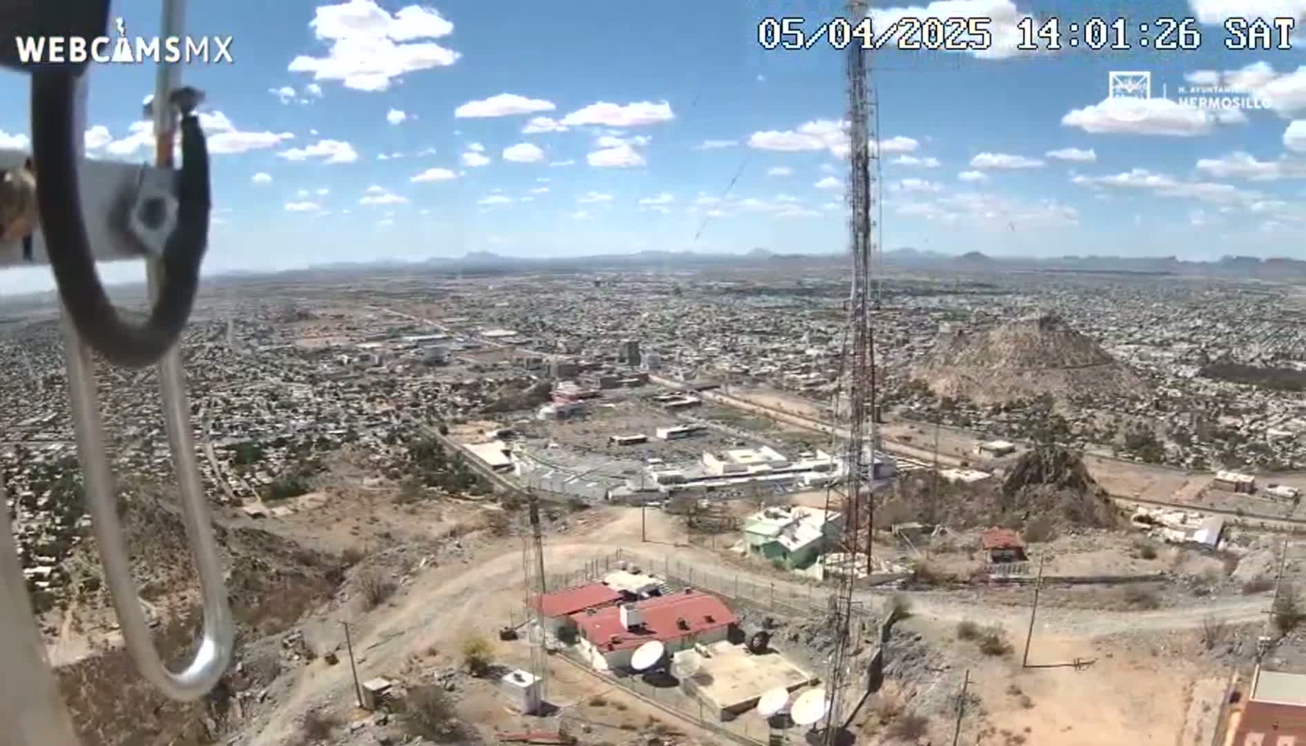 A high-angle, sunny view of a city sprawling across a desert landscape, with a tall communication tower and a building complex on a hill in the foreground.