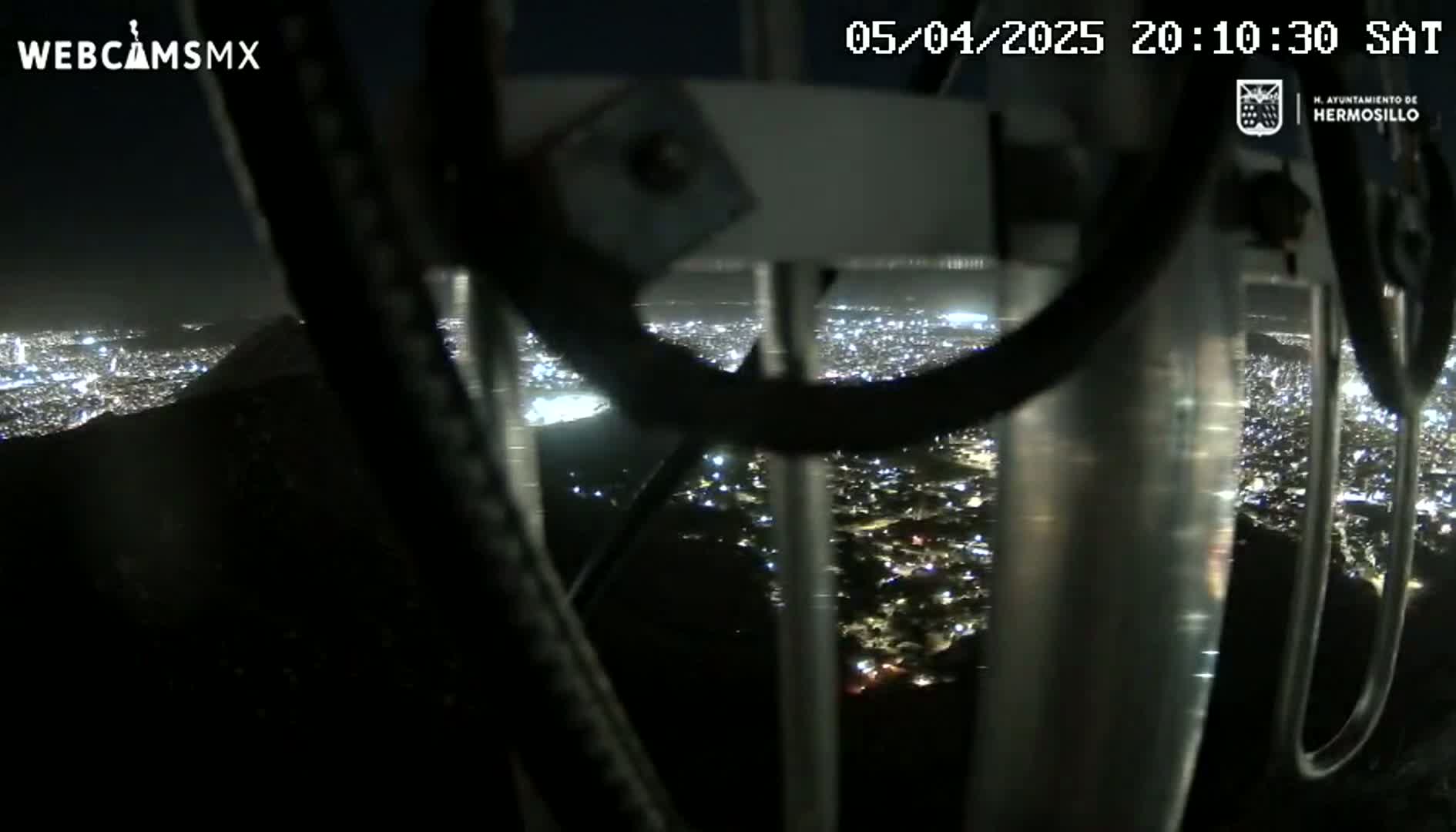A nighttime view of a city illuminated with lights is seen from a high vantage point through the metal bars of a structure.