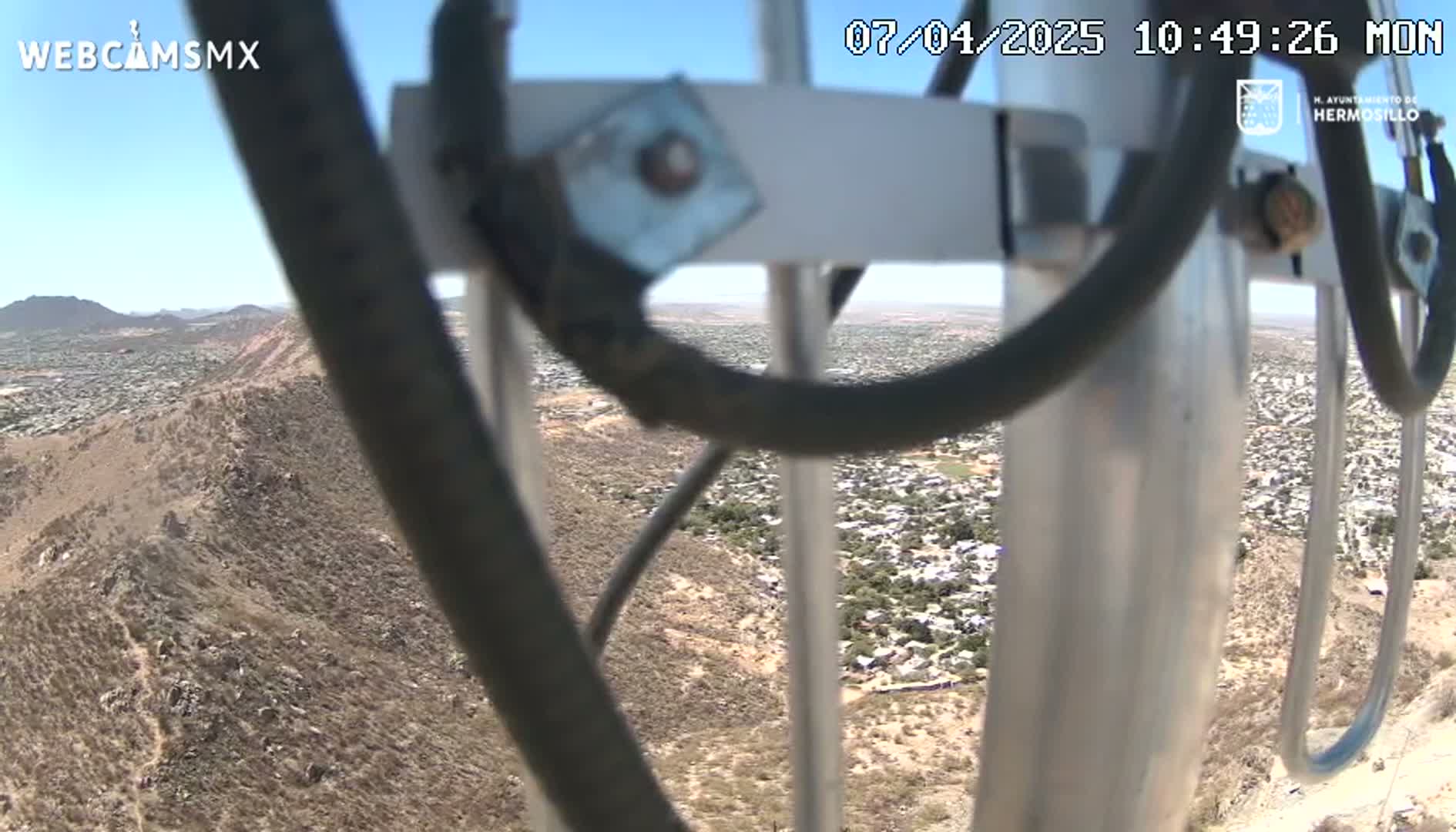 A view of a sprawling town below a rocky, desert hillside on a sunny day, seen through the obstructing wires of a tall pole.