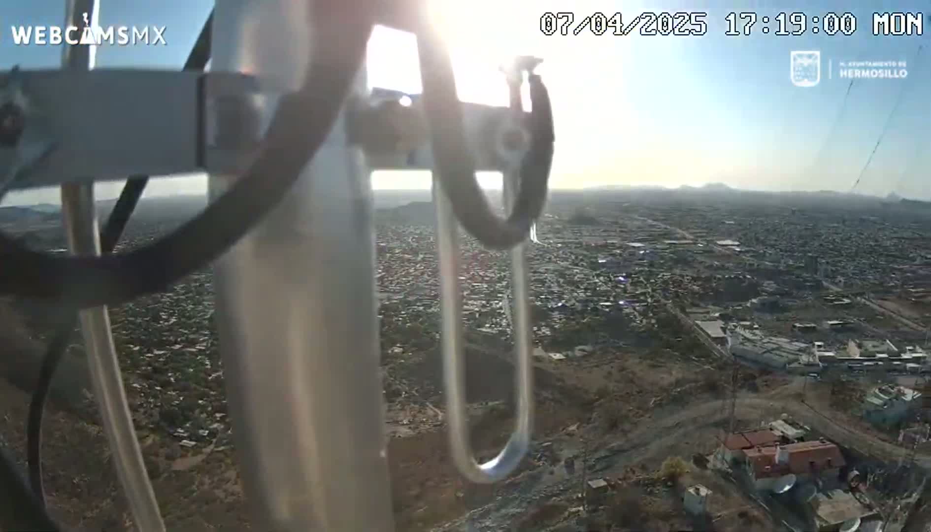 A high-angle, sunlit view from a tall structure shows a sprawling city extending to hazy mountains in the distance on a clear day.