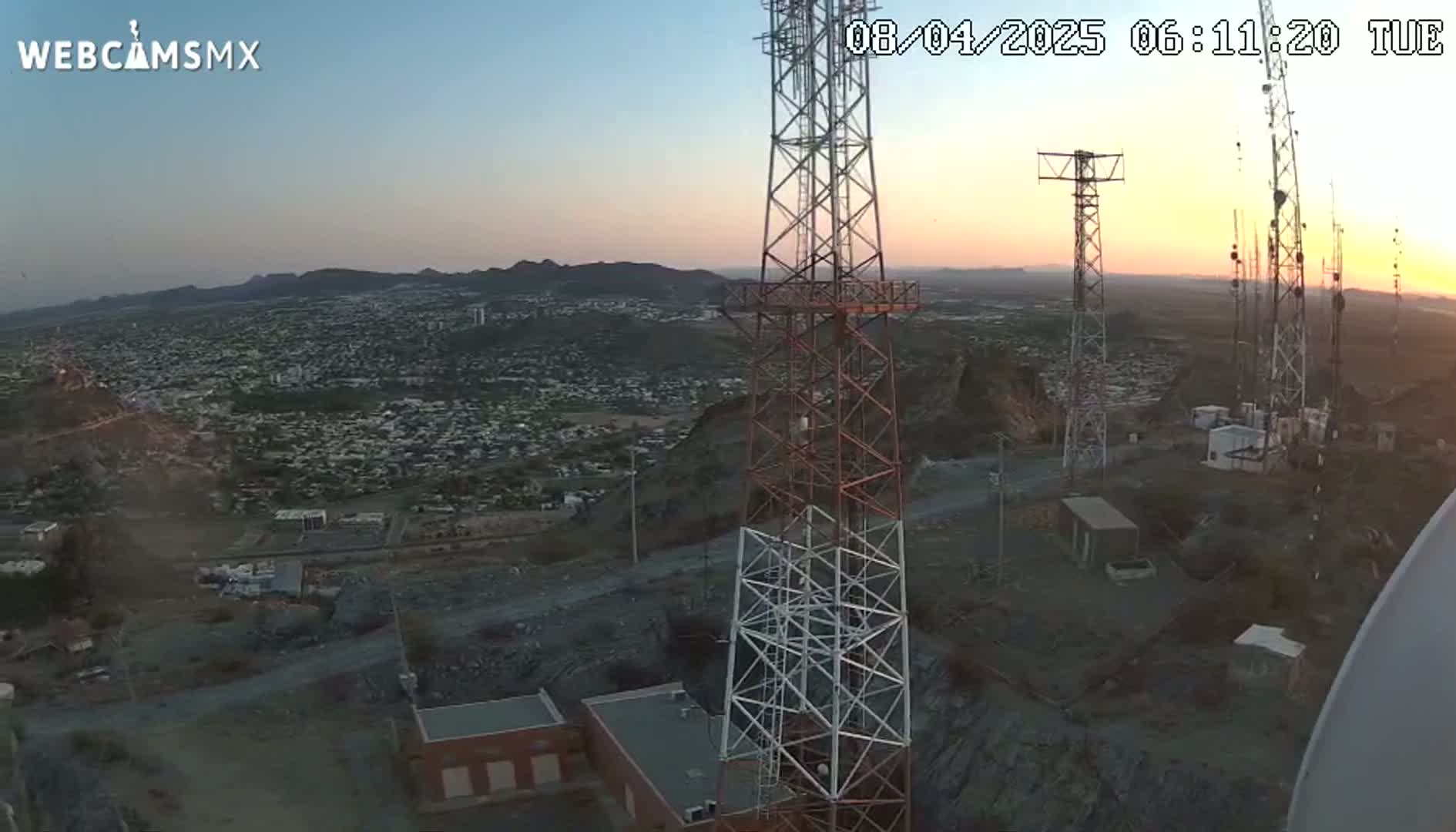 A high-angle, long shot at sunrise shows a sprawling city nestled in a mountainous region, with several communication towers prominently positioned in the foreground.