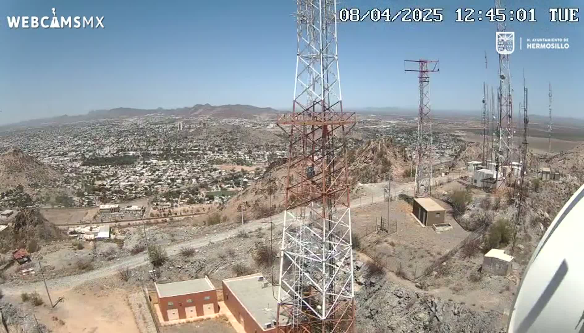 A high-angle view on a clear, sunny day shows a large communication tower atop a rocky hill overlooking a sprawling city and distant mountains.