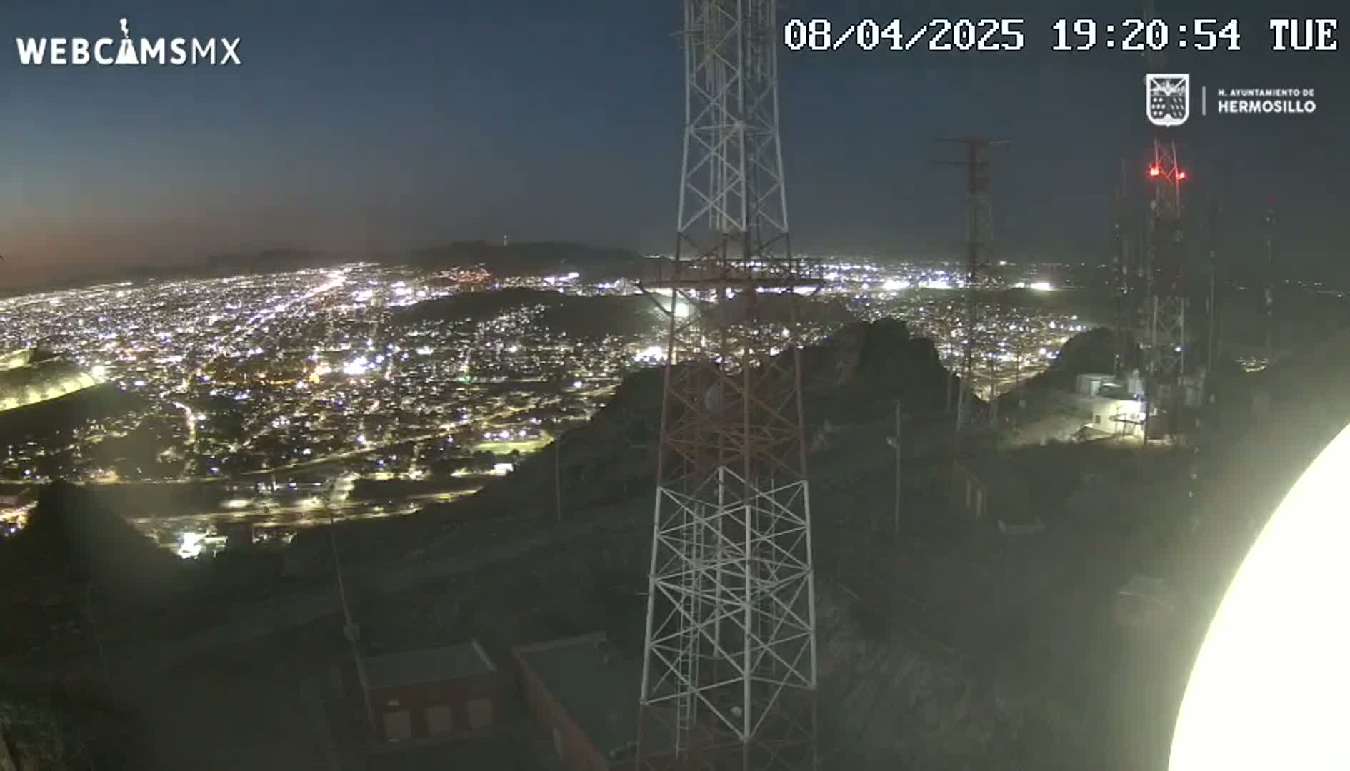 A nighttime view from a hilltop shows a sprawling city illuminated below, with communication towers standing prominently in the foreground under a clear, dark sky.
