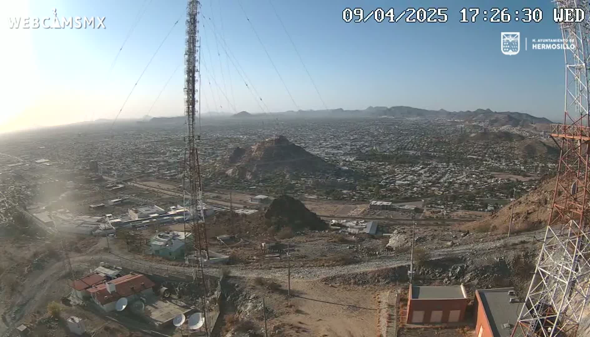 A high-angle, long shot on a clear day shows a sprawling city extending to hazy mountains in the distance, with communication towers and buildings in the foreground.