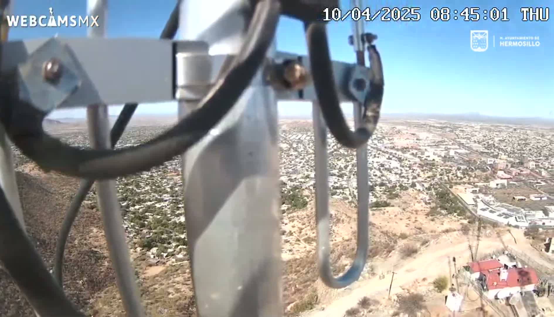 A view from a tall metal pole shows a sprawling city under a clear blue sky on a dry, hilly landscape.