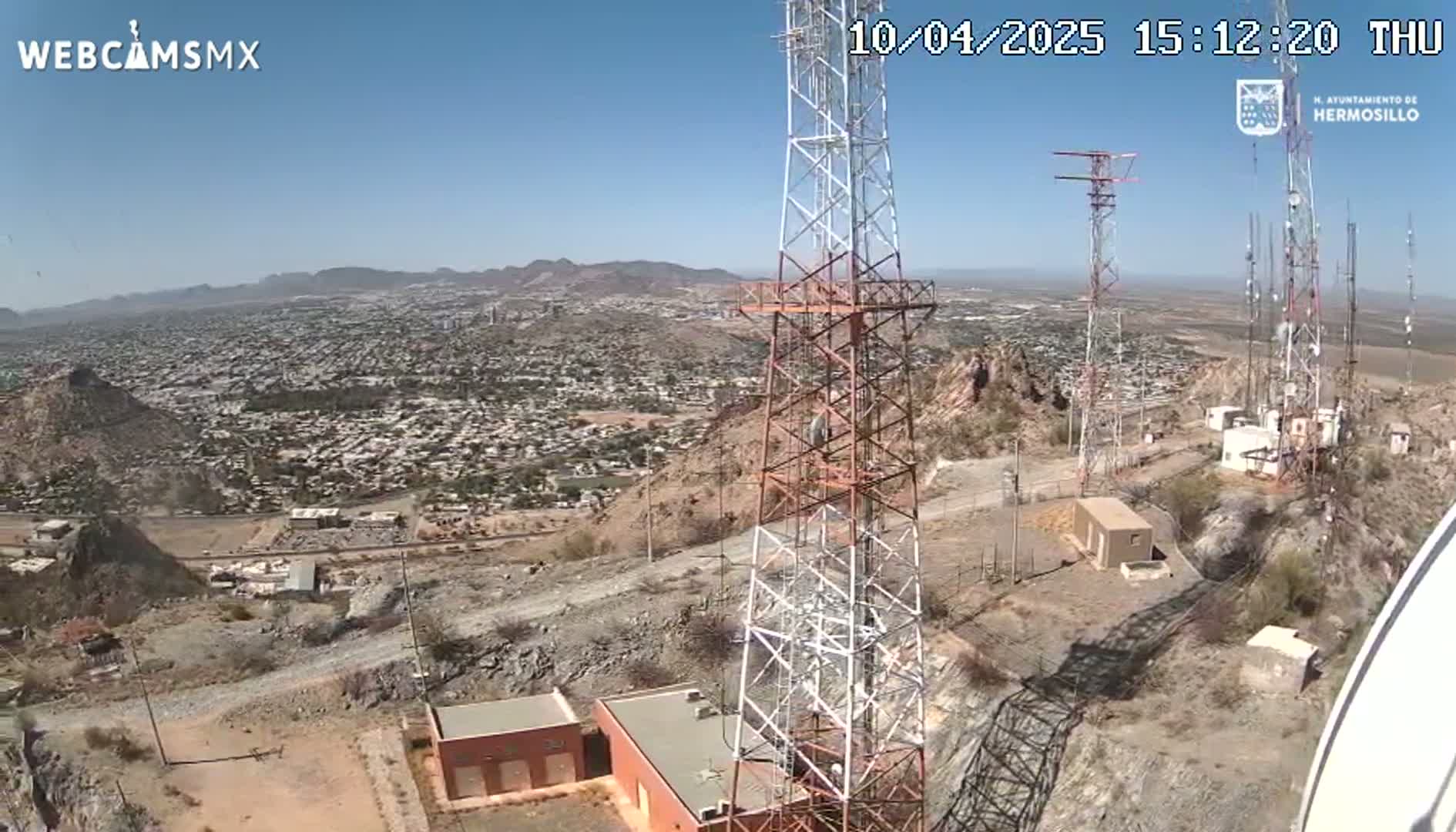A high-angle view on a clear, sunny day shows a sprawling city below several communication towers atop a rocky hill.