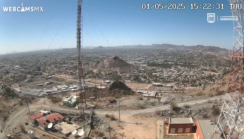 A high-angle, long shot on a clear day shows a large city sprawling across a valley below a rocky hilltop, where communication towers stand amidst sparse structures.