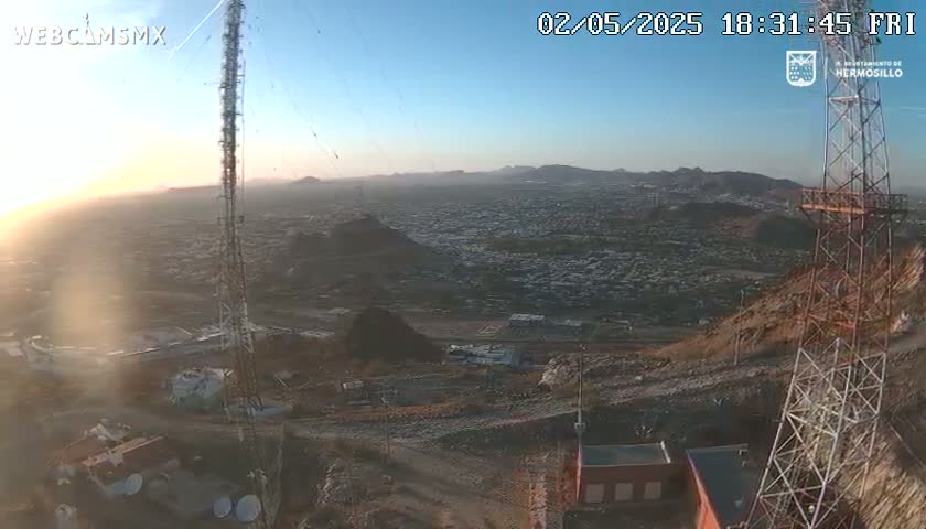 A high-angle, long shot at sunset shows a sprawling city nestled in a mountainous region, with communication towers and structures on a hilltop overlooking the landscape.