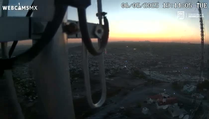 A sunset over a sprawling city is viewed from a high vantage point, partially obscured by the metallic structure of a camera mount.