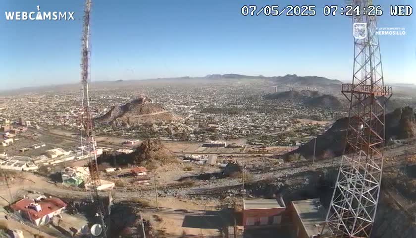 A high-angle, long shot on a clear day shows a sprawling city below a hilltop with communication towers, buildings, and rocky terrain.