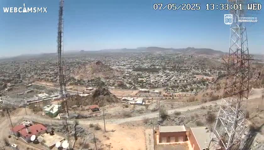 A high-angle, long shot on a clear, sunny day shows a sprawling city nestled in a mountainous region, with several communication towers prominently featured in the foreground.
