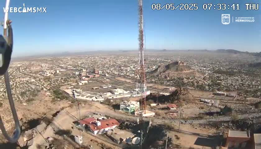 A high-angle, long shot on a clear day shows a city sprawling across a desert landscape, with a tall communication tower and buildings near a rocky hill in the foreground.
