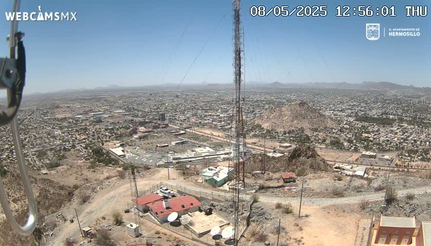 A high-angle, long shot on a sunny day shows a sprawling city in a desert landscape, with a tall communication tower and several buildings situated on a hill overlooking the urban area.