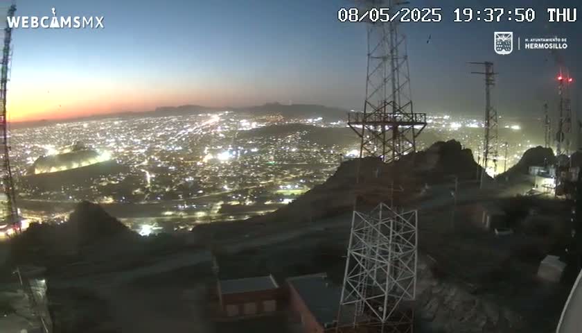 A nighttime, twilight view from a hilltop shows a sprawling city illuminated below, with several tall communication towers nearby.
