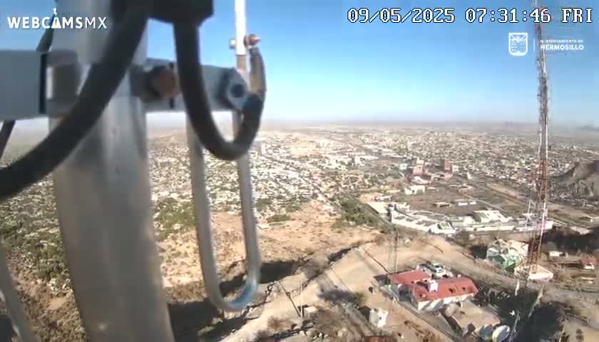 A high-angle view on a clear, sunny day shows a sprawling city extending to the horizon, partially obscured by a tall communication tower and the camera's mounting equipment in the foreground.