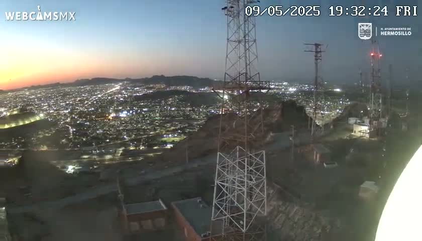 A twilight view from a hilltop overlooking a sprawling city at night, with several tall communication towers in the foreground.