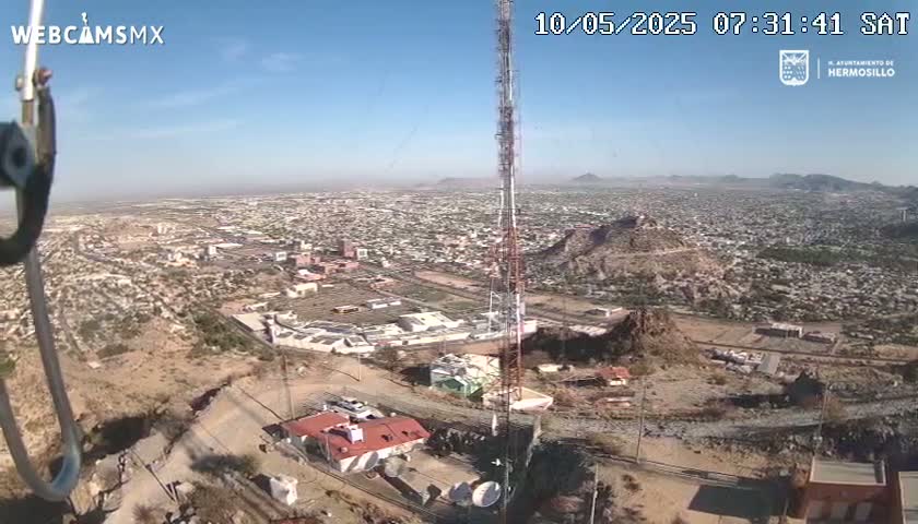 A high-angle, long shot on a clear, sunny day shows a city sprawling across a desert landscape, with a tall communications tower and a building visible in the foreground.