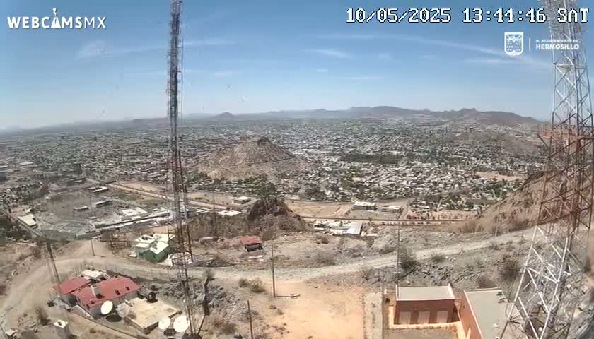 A high-angle, long shot on a clear, sunny day shows a sprawling city nestled in a mountainous region, with communication towers and buildings visible in the foreground.