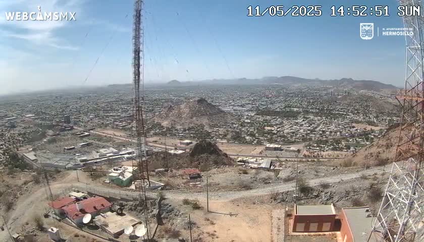 A high-angle view on a sunny day shows a sprawling city below a rocky hilltop with communication towers and several small buildings.