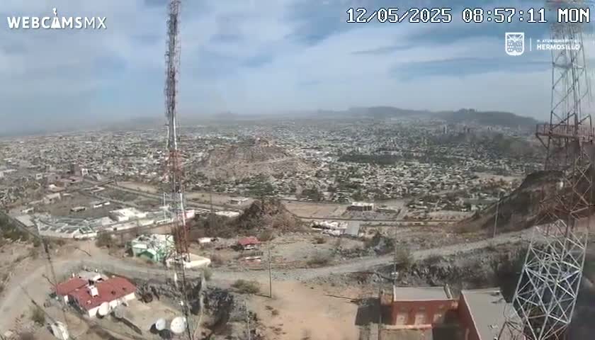 A high-angle view on a partly cloudy day shows a sprawling city below a hilltop with communication towers and several buildings.