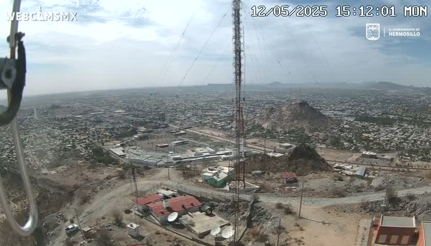 A high-angle view on a partly cloudy day shows a large city sprawling across a valley, with a tall communications tower and several buildings situated on a rocky hillside overlooking it.