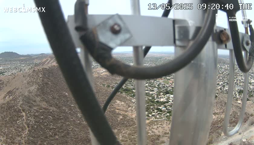 A view of a sprawling town nestled in a mountainous, arid landscape is seen through the metal framework of a tall structure on a partly cloudy day.