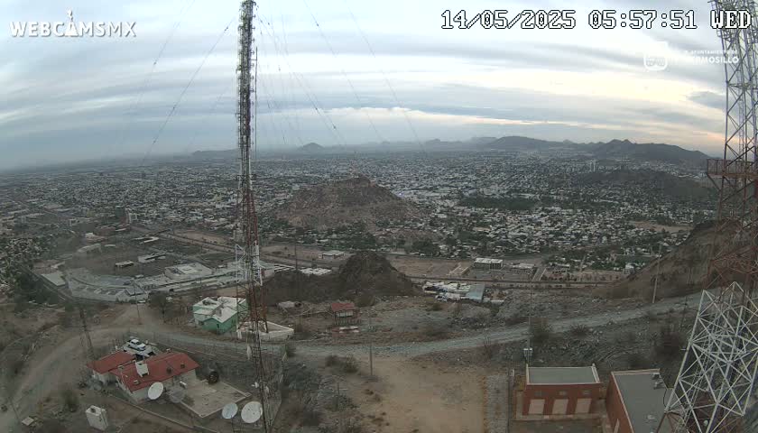 A large city sprawls beneath an overcast sky, viewed from a hilltop where communication towers stand.