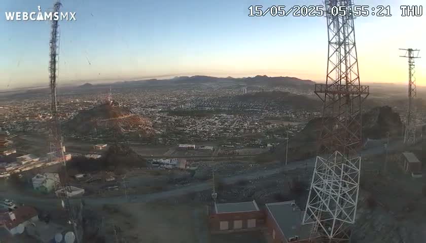 A sunrise view from a hilltop shows a sprawling city below, with communication towers in the foreground and clear skies.