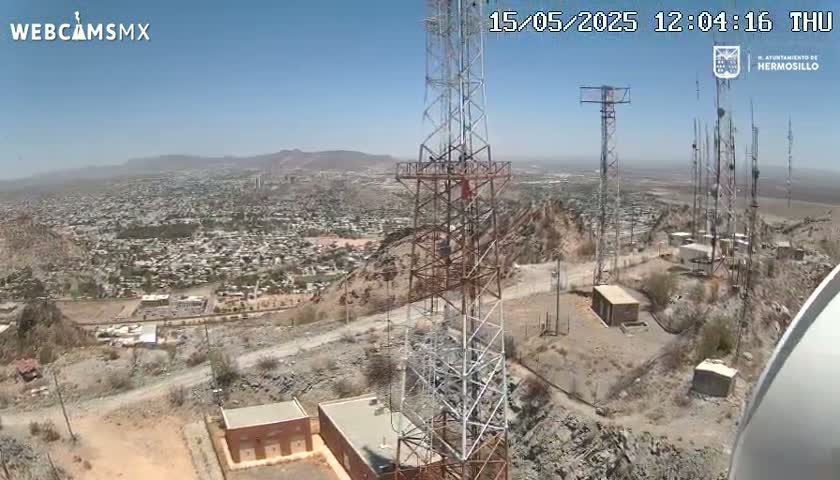 A high-angle view on a clear, sunny day shows several communication towers atop a rocky hill overlooking a sprawling city and distant mountains.