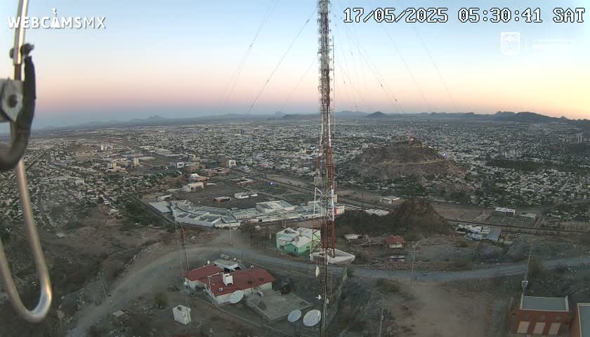 An elevated view at dawn shows a large city sprawling across a valley, with a tall communication tower and several buildings situated on a rocky hill overlooking the urban landscape.